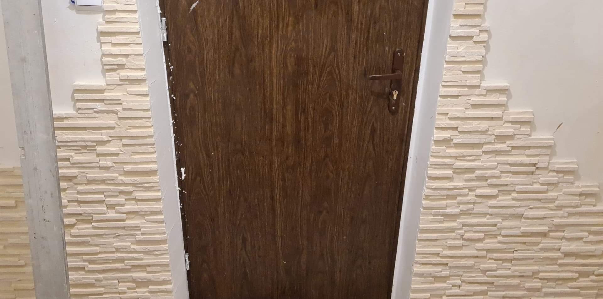 Ciemnobrązowe drzwi z brązową klamką, obok ściana wykończona dekoracyjną, beżową okładziną imitującą kamień.