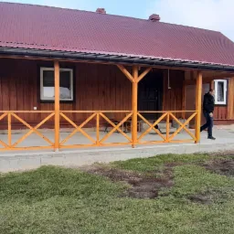 Drewniany dom z nowym, pomalowanym na brązowo dachem z blachy, jasną, drewnianą werandą z kratownicą i przechodzącym mężczyzną w czarnej kurtce.