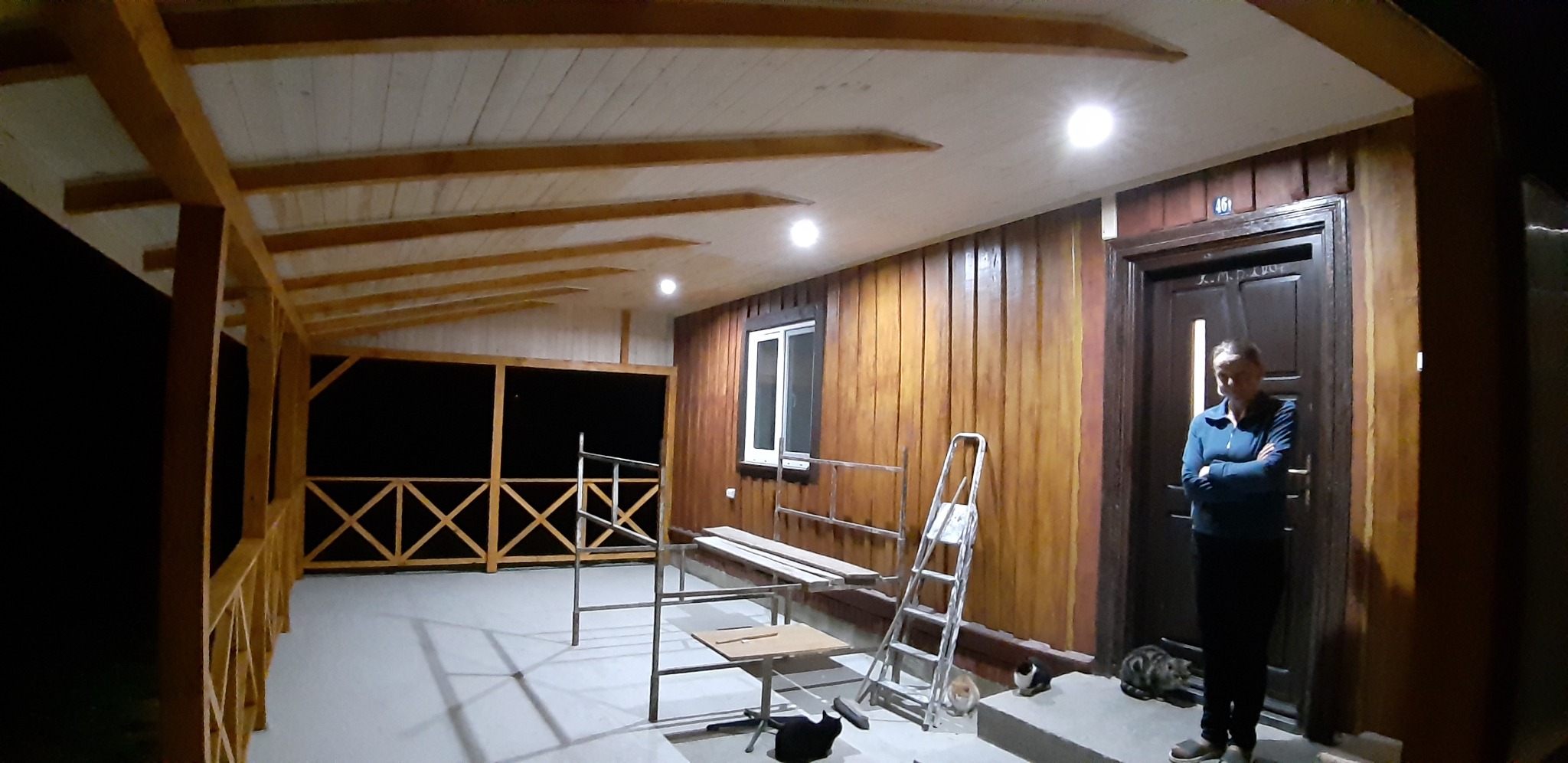 Zadaszony taras z drewnianą konstrukcją i podbitką, oświetlony punktowymi lampami, z elementami rusztowania, drabiną i osobą stojącą przy drzwiach wejściowych.