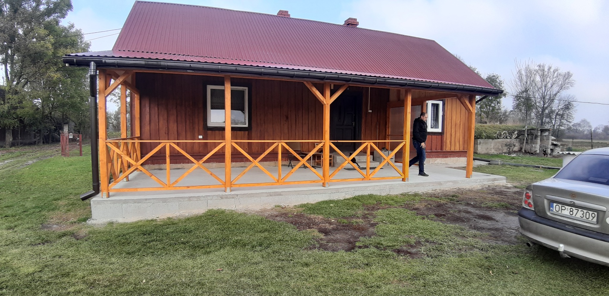 Drewniany dom z nowym, pomalowanym na brązowo dachem z blachy, jasną, drewnianą werandą z kratownicą i przechodzącym mężczyzną w czarnej kurtce.