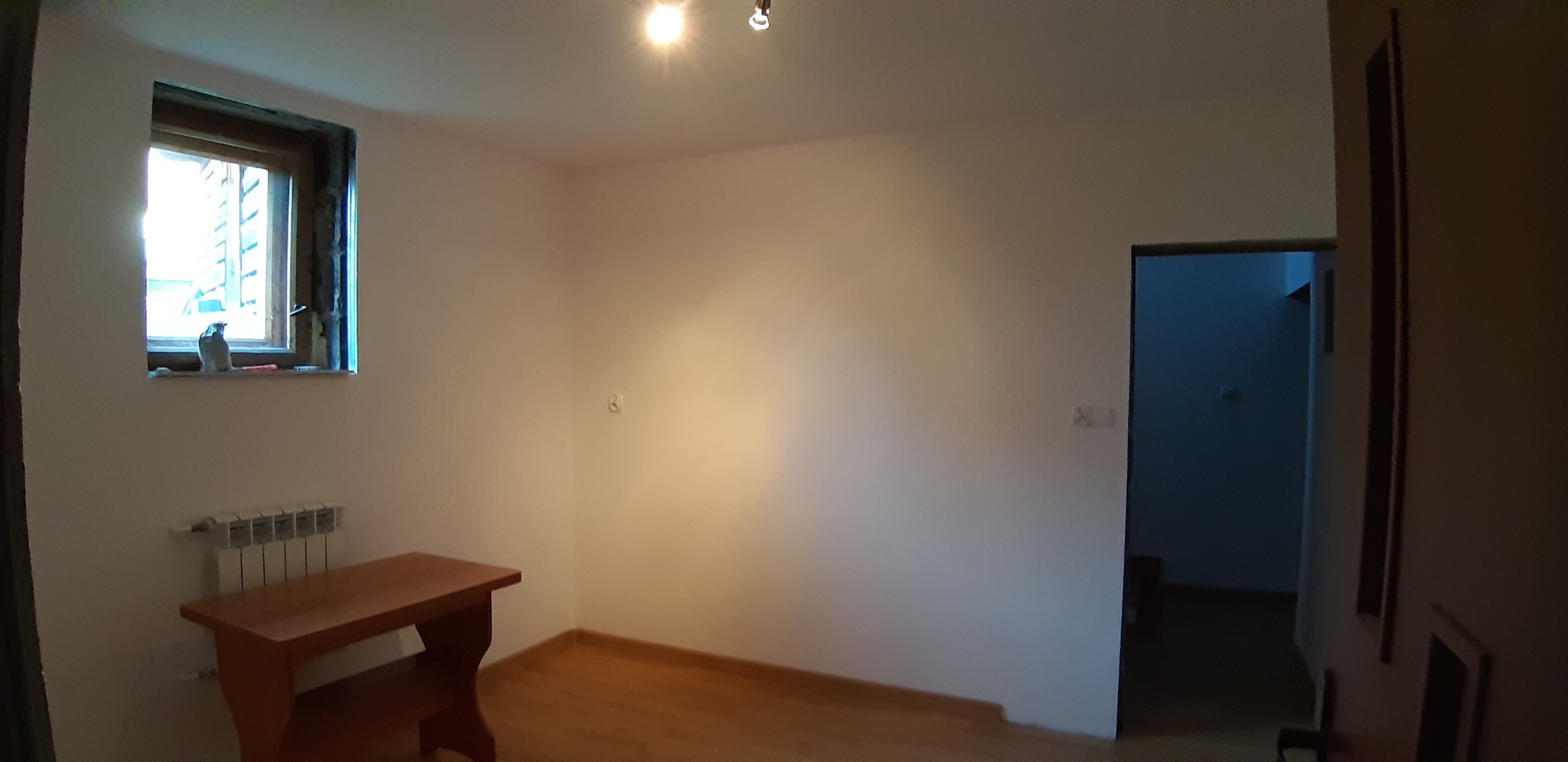 Pusty pokój z drewnianą podłogą, grzejnikiem pod oknem i drewnianym stolikiem, przygotowany do prac wykończeniowych, z widocznym oświetleniem sufitowym i otwartymi drzwiami do kolejnego pomieszczenia.