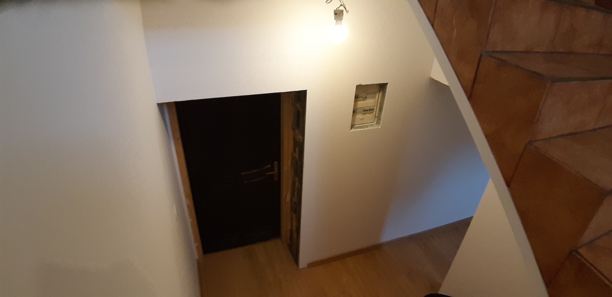 Wnętrze klatki schodowej z widocznymi schodami z terakoty, ścianami po malowaniu oraz osadzoną w ścianie skrzynką elektryczną i drzwiami z ościeżnicą w trakcie montażu.