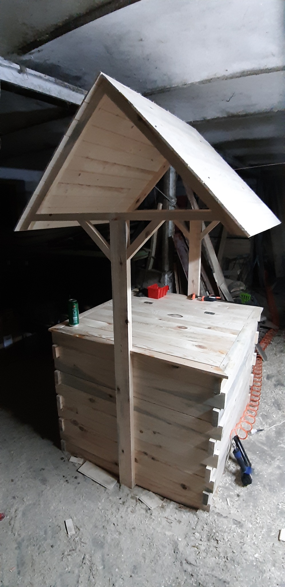 Drewniana studnia ogrodowa w trakcie budowy, widoczne narzędzia i elementy konstrukcyjne, surowe drewno, betonowy strop w tle.