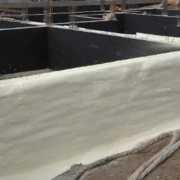 Fundamenty budynku po izolacji natryskowej pianką, widoczne pionowe pręty zbrojeniowe i czarna hydroizolacja, ziemia wokół fundamentów.