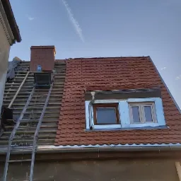 Częściowo zdjęty dach z widoczną konstrukcją drewnianą, komin z cegły, drabina oparta o budynek, nowe dachówki na części dachu, okno dachowe z obróbką blacharską i rynny. Budynek z widocznymi...