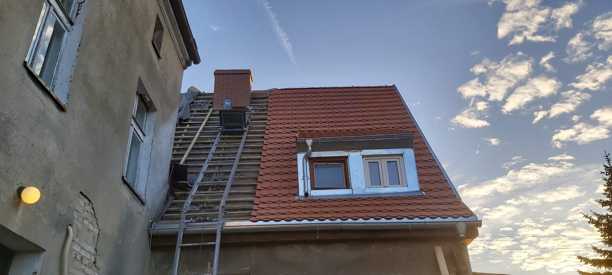 Częściowo zdjęty dach z widoczną konstrukcją drewnianą, komin z cegły, drabina oparta o budynek, nowe dachówki na części dachu, okno dachowe z obróbką blacharską i rynny. Budynek z widocznymi...