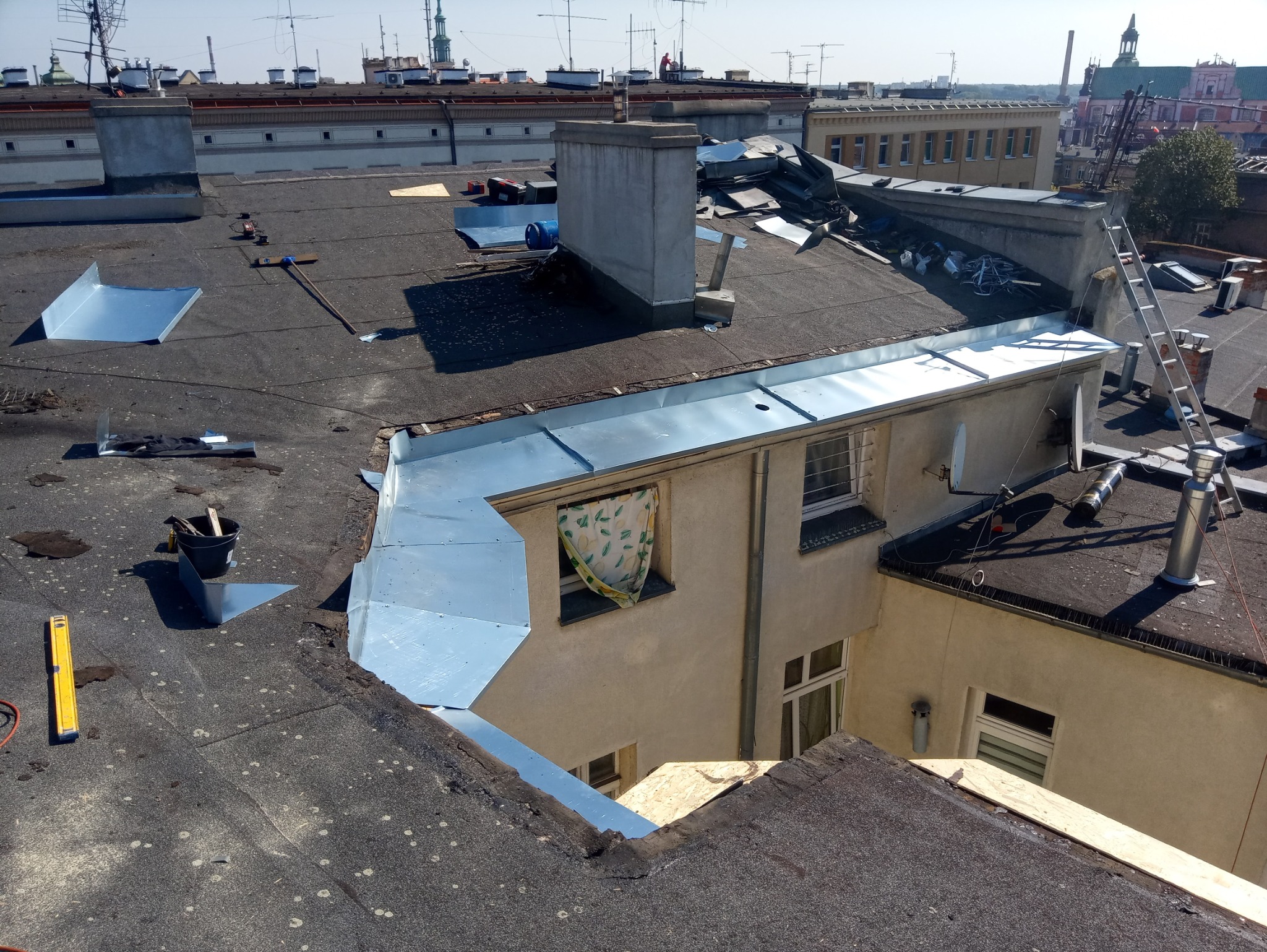 Widok z góry na dach budynku w trakcie prac blacharskich, z widocznymi nowymi elementami obróbek blacharskich, narzędziami, wiadrem i poziomicą, w tle zabudowa miejska.