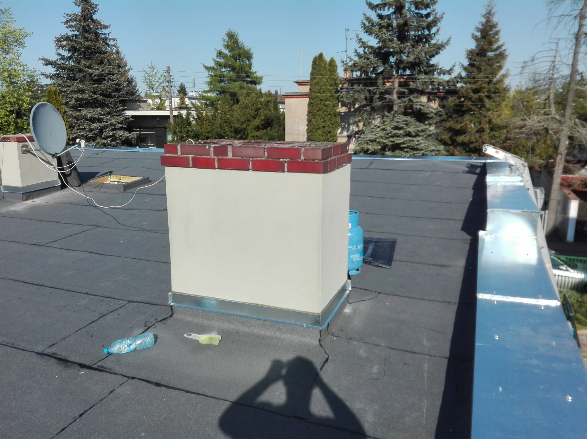 Płaski dach pokryty papą z kominem obłożonym cegłą klinkierową, widoczna butla gazowa, butelka wody i szczotka do czyszczenia, w tle drzewa i budynki.