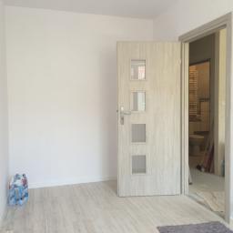 PHU Piotr Kuczyński - Wnętrze pokoju z otwartymi drzwiami do wyremontowanej łazienki, widoczne białe ściany, jasna podłoga i butelki wody mineralnej w rogu.