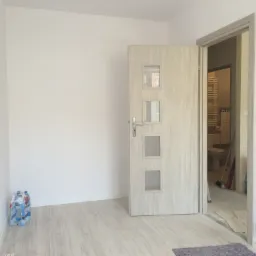 Wnętrze pokoju z otwartymi drzwiami do wyremontowanej łazienki, widoczne białe ściany, jasna podłoga i butelki wody mineralnej w rogu.