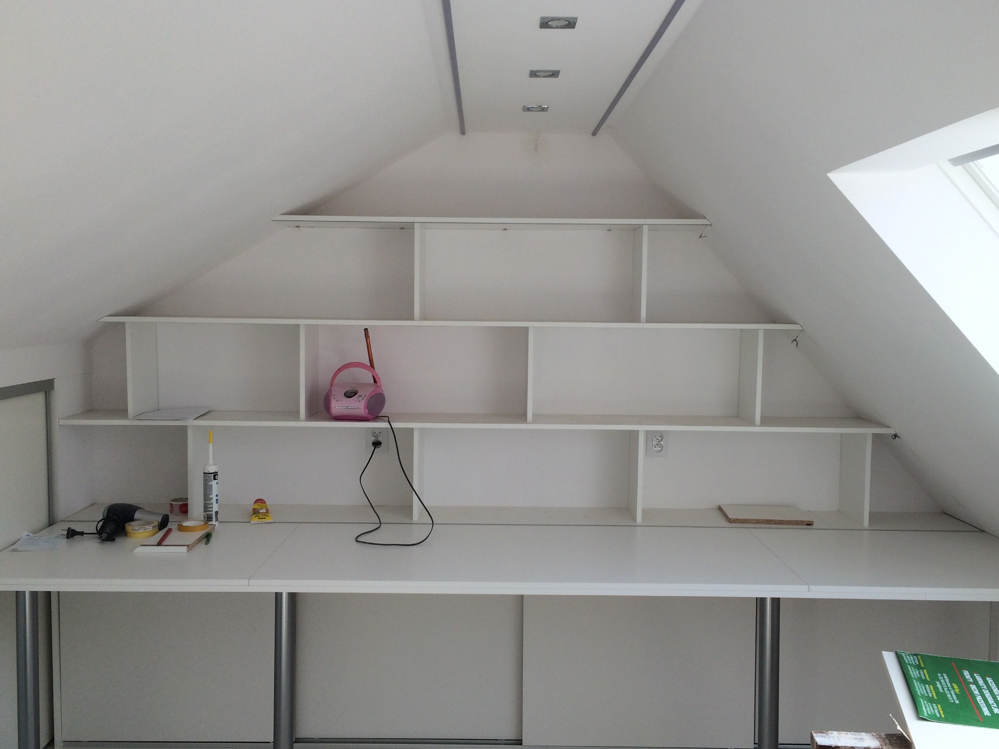 Białe, niestandardowe półki wbudowane w skos dachu, z blatem roboczym poniżej, widoczne narzędzia i akcesoria na blacie, gniazdka elektryczne na ścianie.