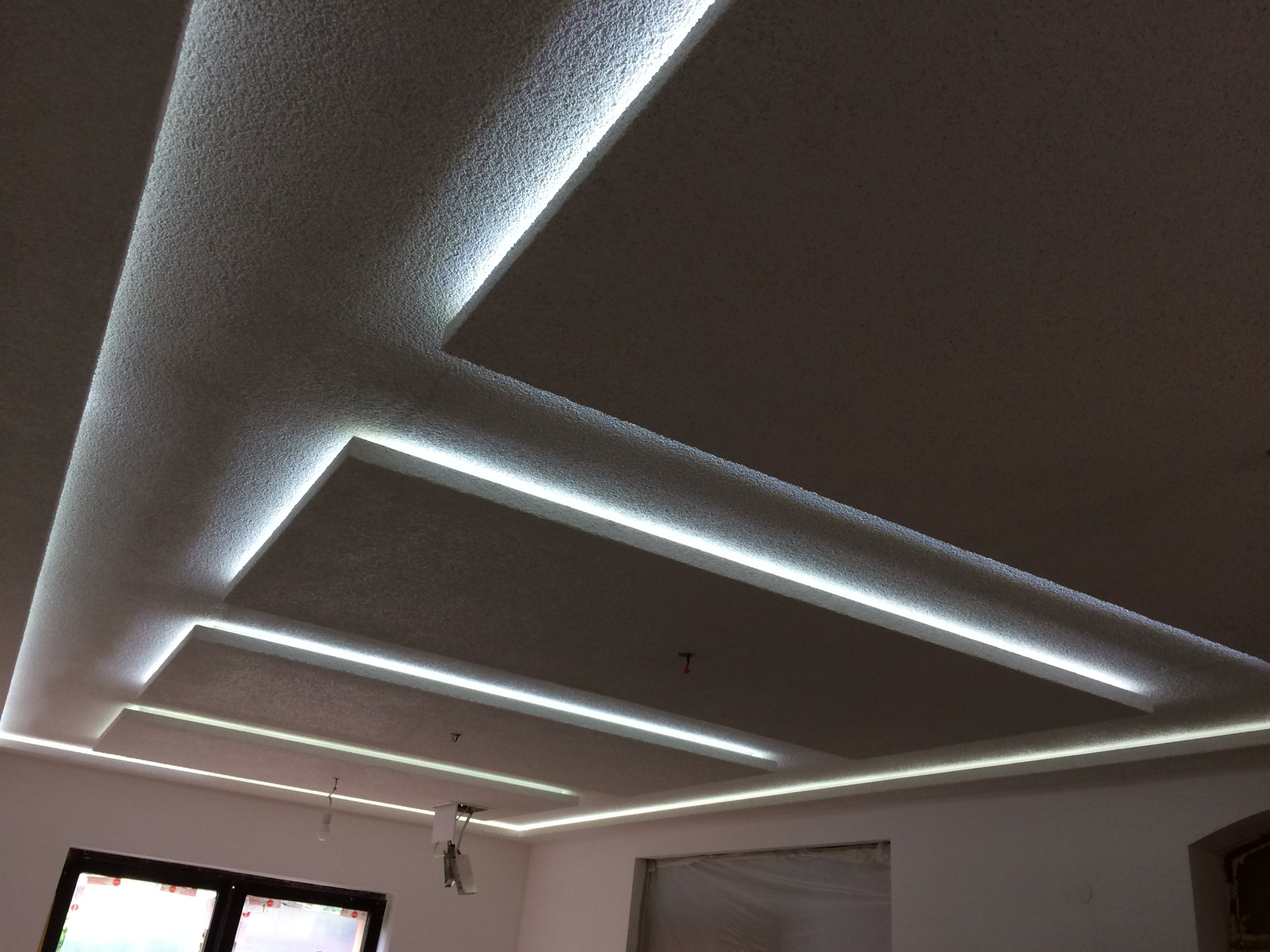 Kaskadowy sufit podwieszany z wbudowanym oświetleniem LED, wykończony gładzią strukturalną, widoczne okno i fragment ściany w trakcie prac wykończeniowych.