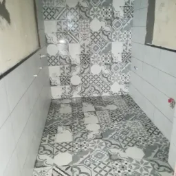 Ukończona łazienka z szaro-białymi płytkami o wzorach geometrycznych i ornamentacyjnych na podłodze i ścianach; widoczne wystające rurki instalacji wodnej.