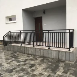 Czarna, metalowa balustrada przy wejściu do domu z szarą elewacją i kostką brukową na podjeździe, widoczne dwa okna na fasadzie.