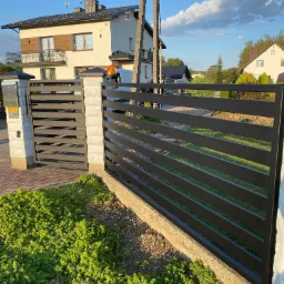 Nowoczesna, pozioma brama wjazdowa z metalu w kolorze antracytu, z furtką po lewej stronie, na tle domu jednorodzinnego i zieleni.