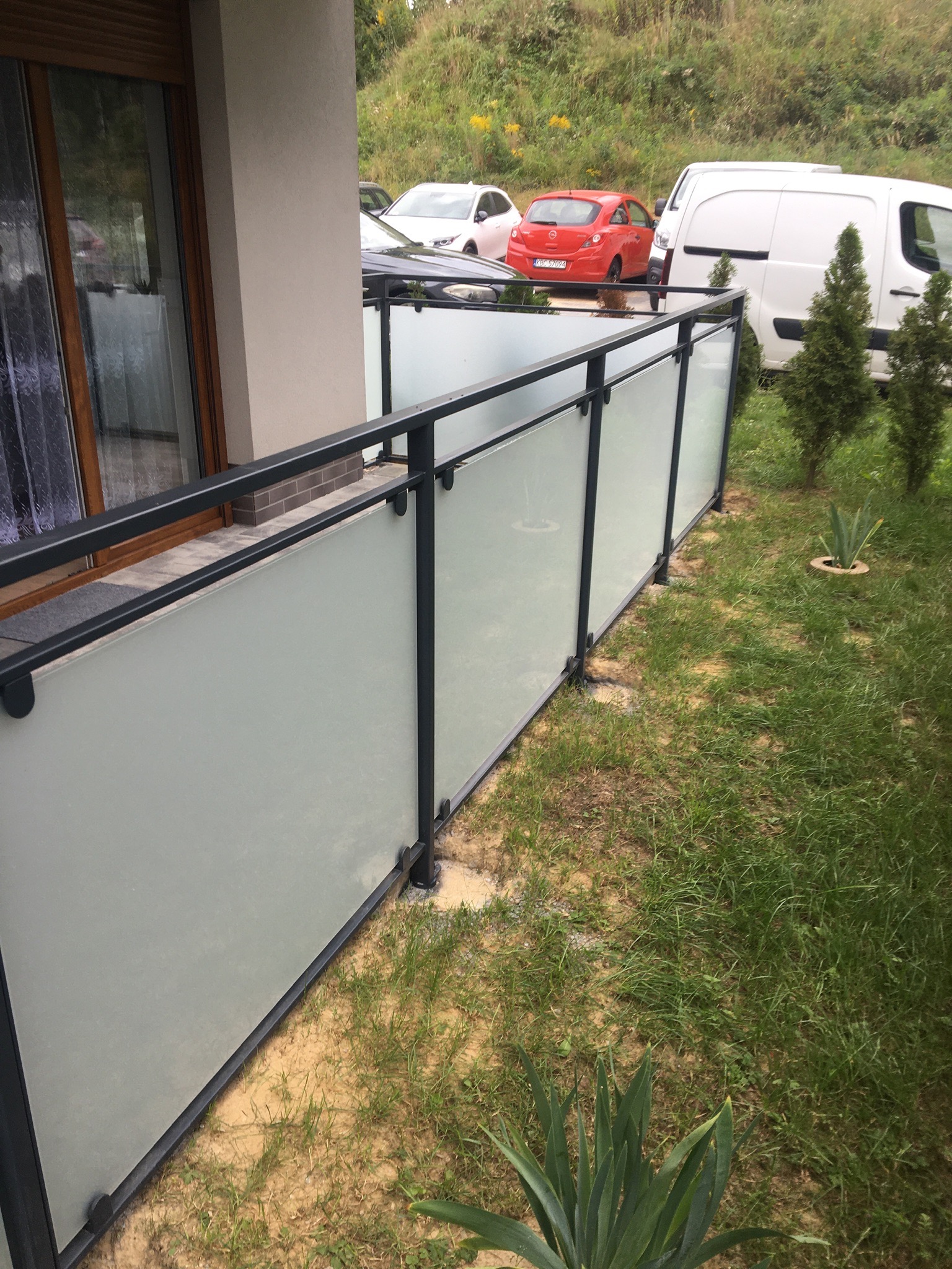 Nowoczesna balustrada z matowego szkła i czarnych profili, zamontowana przy tarasie domu, widok z boku na ogród i zaparkowane samochody w tle.