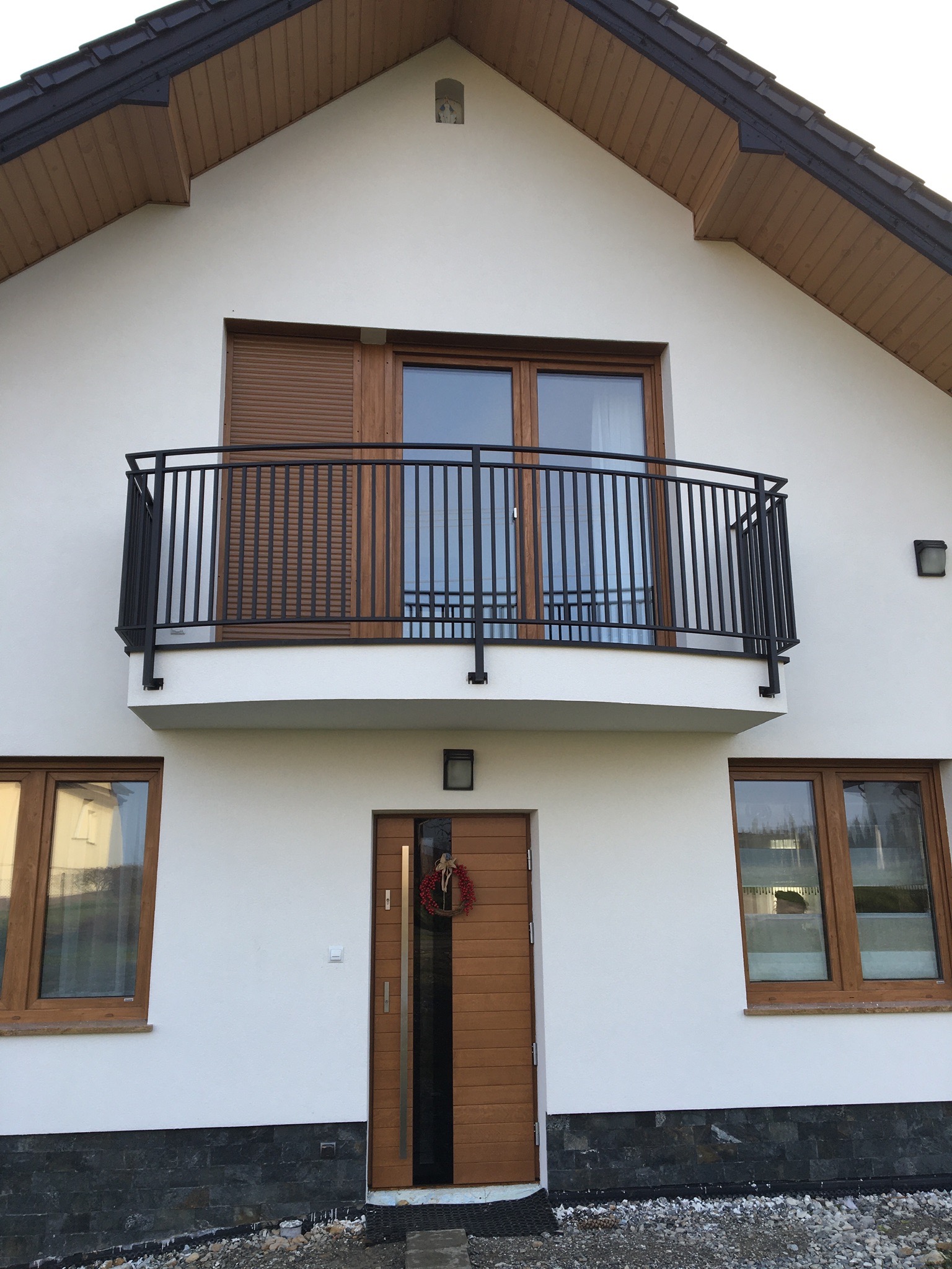 Biały dom z czarną, metalową balustradą balkonową, drewnianymi oknami i drzwiami wejściowymi z wieńcem świątecznym.