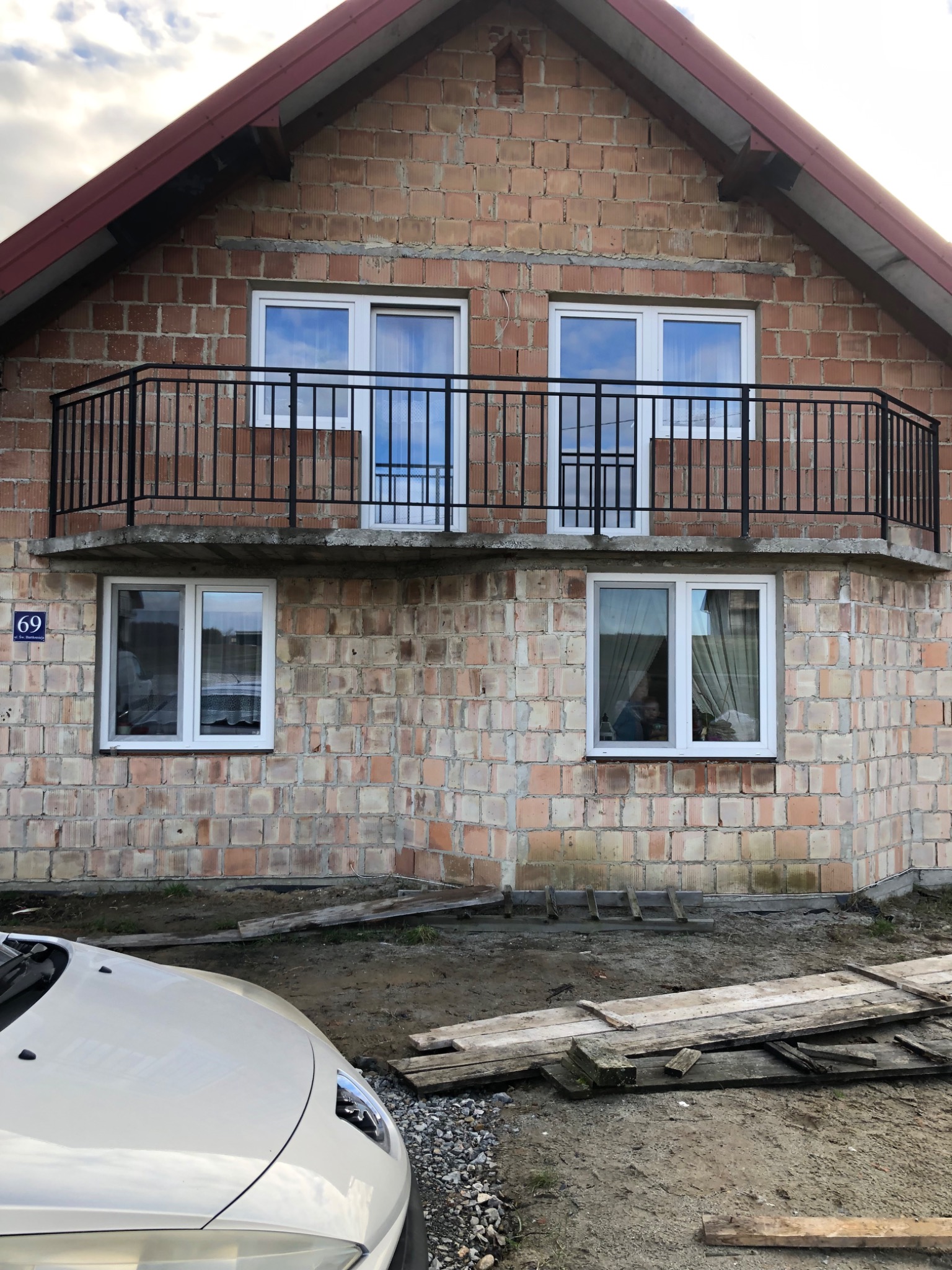 Surowy stan domu w budowie z czarną, metalową balustradą balkonową i widocznym numerem domu na elewacji.