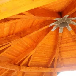 Wnętrze drewnianej altany z widoczną konstrukcją dachu, centralnym metalowym łącznikiem i jasnym oświetleniem.