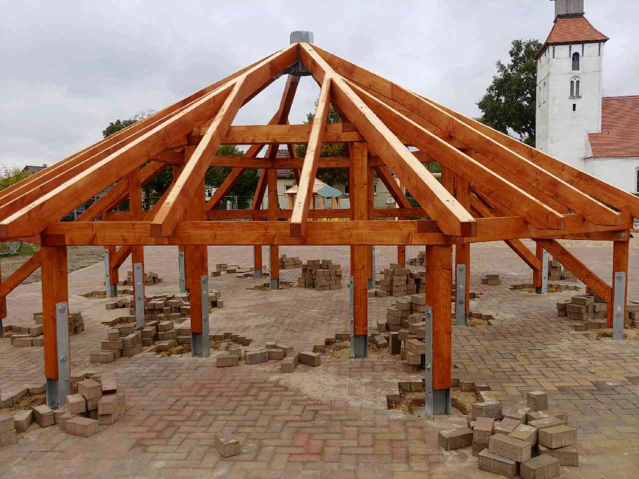 Drewniana konstrukcja z belek przygotowana pod zadaszenie, widoczne metalowe mocowania słupów, kostka brukowa wokół w trakcie układania, w tle budynek z wieżą.