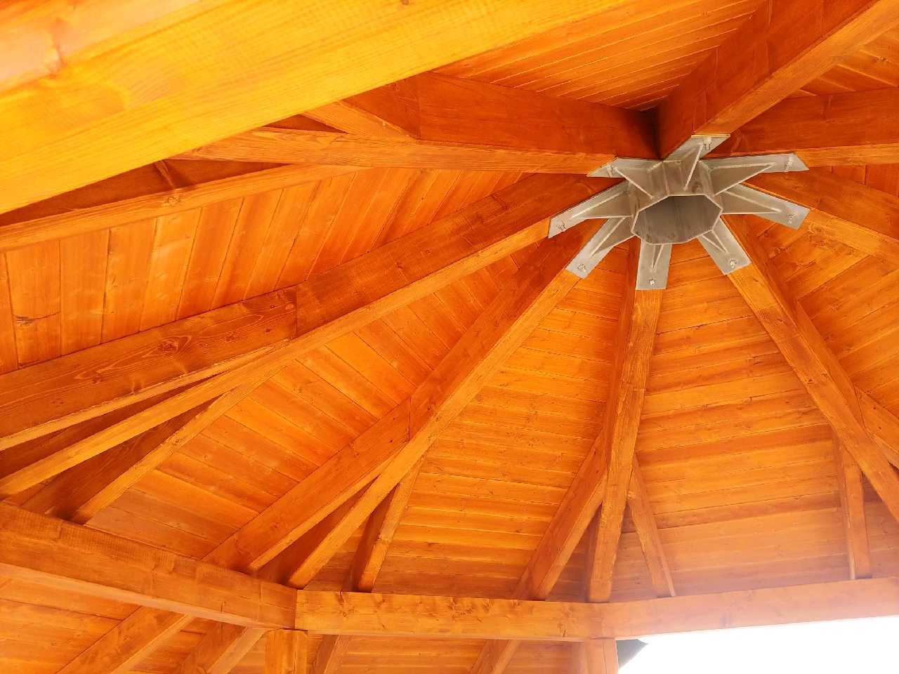 Wnętrze drewnianej altany z widoczną konstrukcją dachu, centralnym metalowym łącznikiem i jasnym oświetleniem.