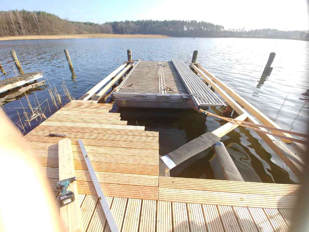 Budowa drewnianego pomostu nad jeziorem, widoczne narzędzia i elementy konstrukcyjne, częściowo ukończona platforma.