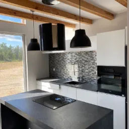 Jasne wnętrze kuchni z wyspą, drewnianymi belkami na suficie i nowoczesnym oświetleniem; widok na zewnątrz przez duże okno.