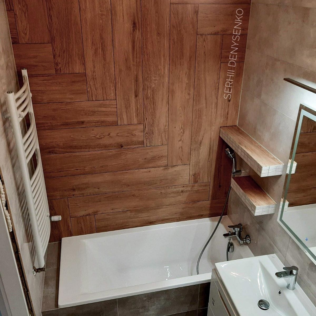 Nowoczesna łazienka z drewnopodobnymi płytkami ułożonymi w jodełkę na ścianie, białą wanną i grzejnikiem drabinkowym, oraz półkami z mozaiką nad baterią wannową.