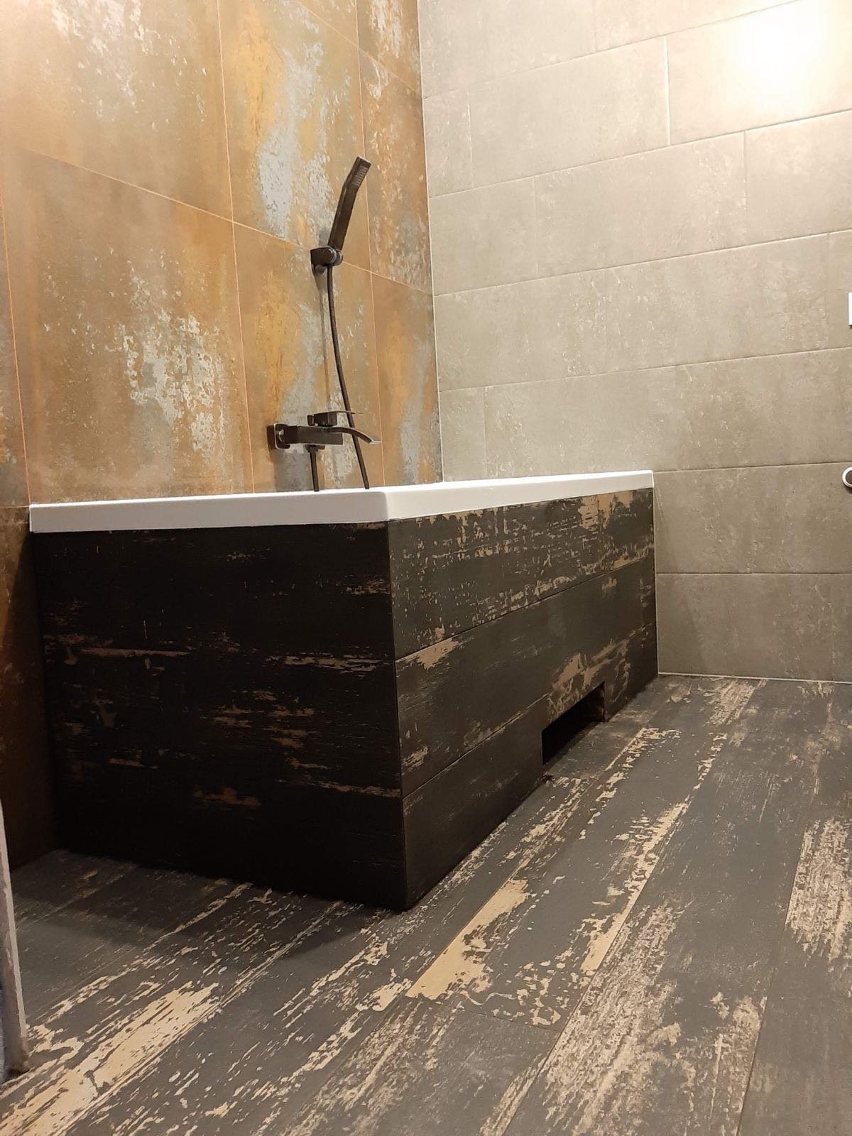 Rustykalna łazienka z wanną obudowaną ciemnym drewnem z efektem postarzenia, płytki imitujące rdzę na ścianie, szara podłoga w stylu vintage.