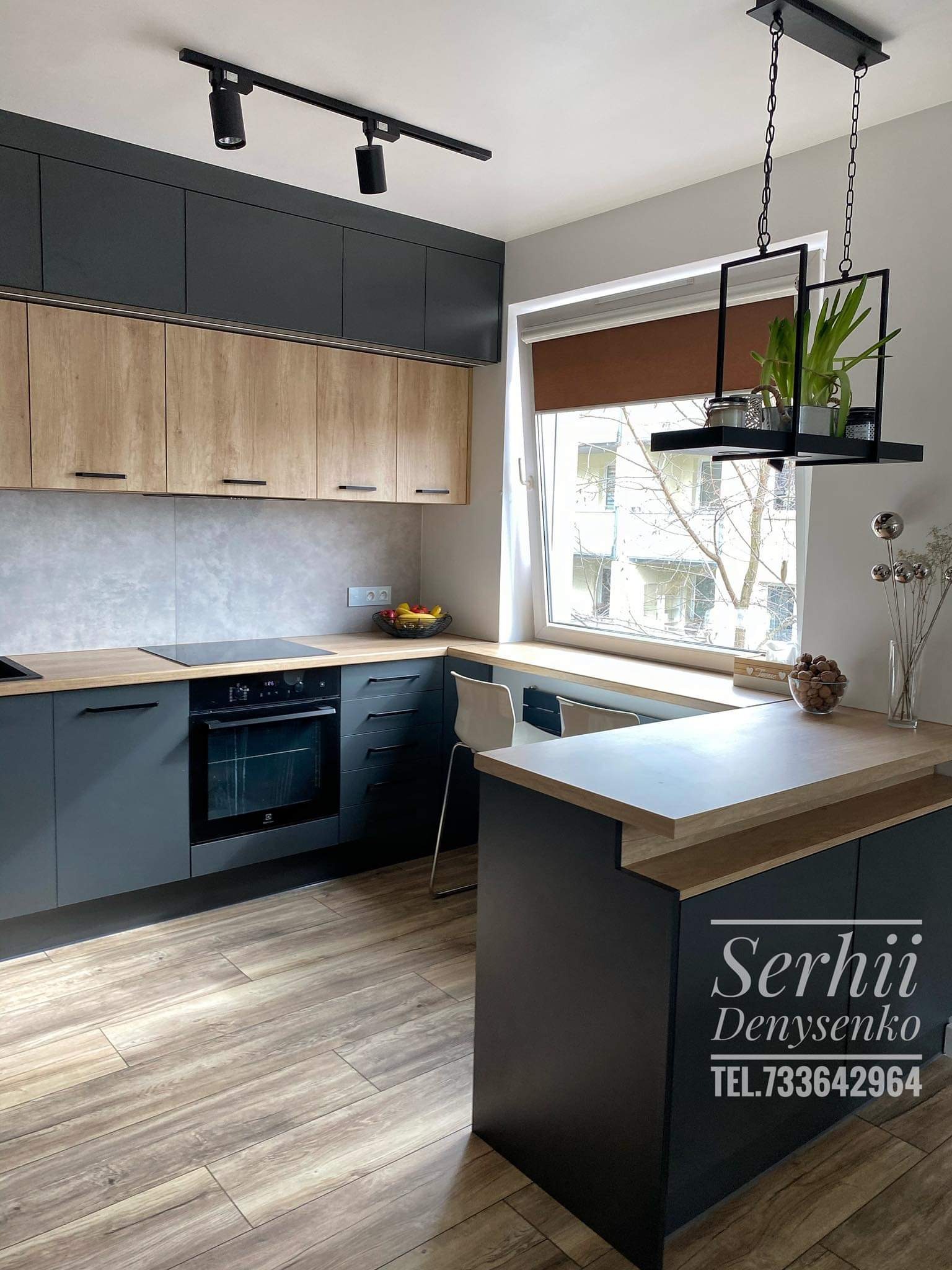 Nowoczesna kuchnia z szarymi szafkami dolnymi i drewnopodobnymi górnymi, drewnianym blatem, czarną płytą indukcyjną i piekarnikiem, oświetleniem szynowym oraz wiszącą dekoracją z roślinami.