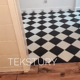 Ułożona podłoga w szachownicę z czarno-białych płytek ceramicznych, widoczny fragment drewnianej podłogi i ramy drzwiowej.