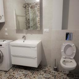 Remonty mieszkań Wrocław - Nowoczesna łazienka po remoncie z białą szafką pod umywalką, toaletą z chromowanym przyciskiem spłukiwania i wzorzystą podłogą z płytek.