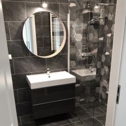 Remonty mieszkań Wrocław - Nowoczesna łazienka w odcieniach szarości z heksagonalnymi płytkami pod prysznicem, okrągłe lustro z podświetleniem nad białą umywalką z czarną szafką, widok zza uchylonych drzwi.