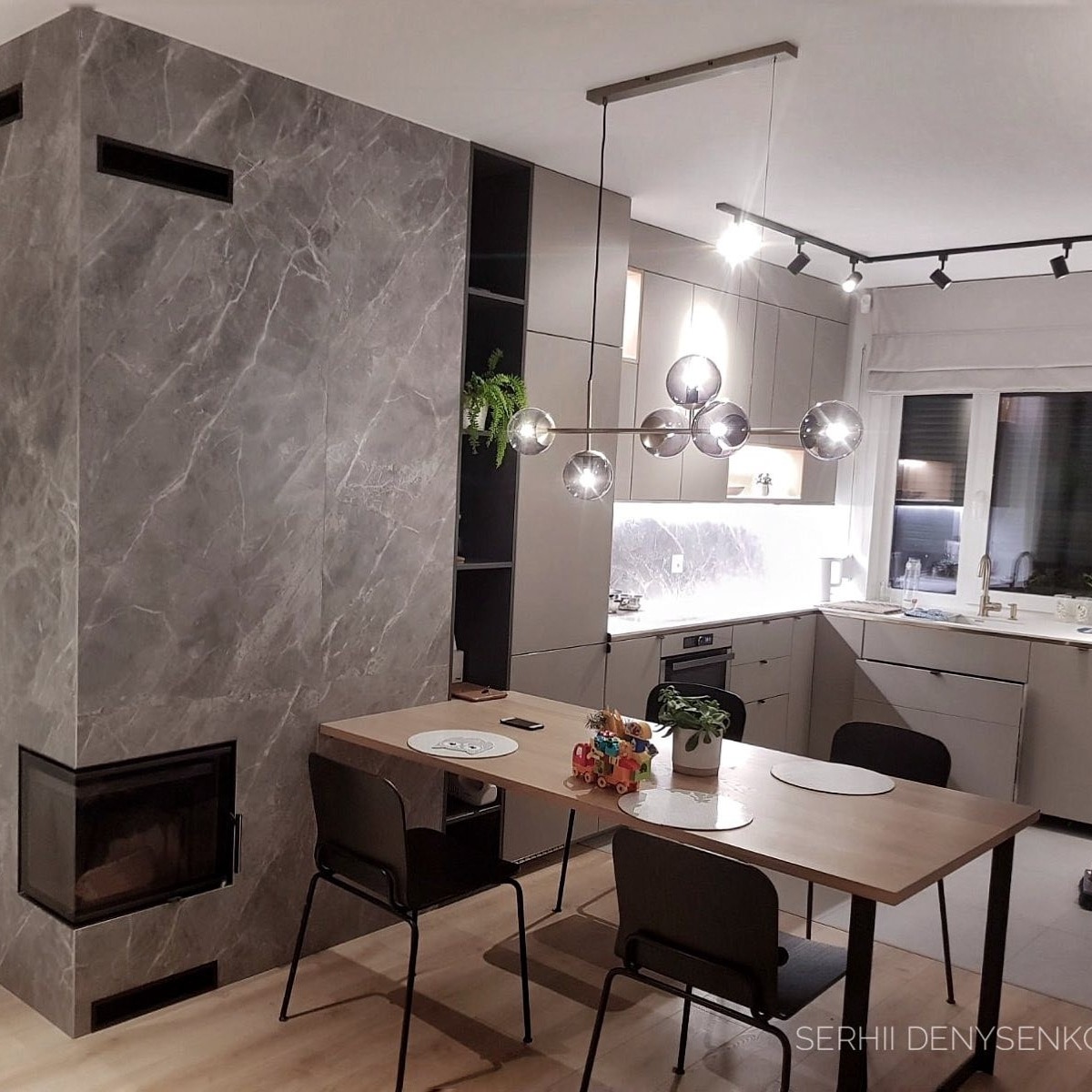 Nowoczesna kuchnia z szarymi szafkami, marmurowym kominkiem narożnym i drewnianym stołem z czarnymi krzesłami, oświetlona kulistą lampą wiszącą i reflektorami.