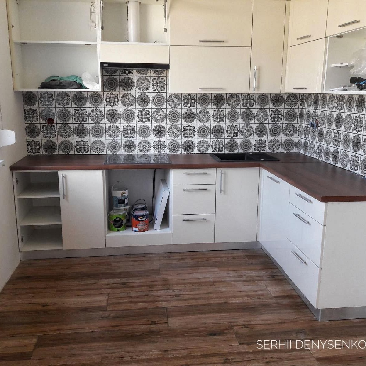 Biała kuchnia z drewnianym blatem i ozdobnymi płytkami na ścianie, widoczne puszki z farbą Dulux w otwartej szafce, podłoga z paneli imitujących drewno.