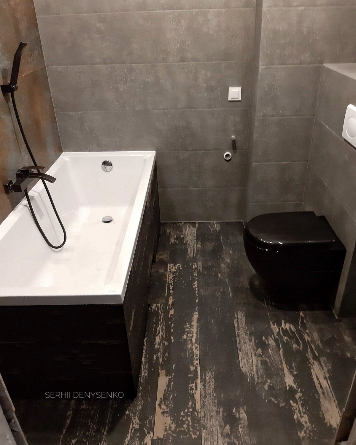 Nowoczesna łazienka z wanną obudowaną ciemnym drewnem i czarną toaletą, szare płytki na ścianach i podłodze z efektem postarzanego drewna.