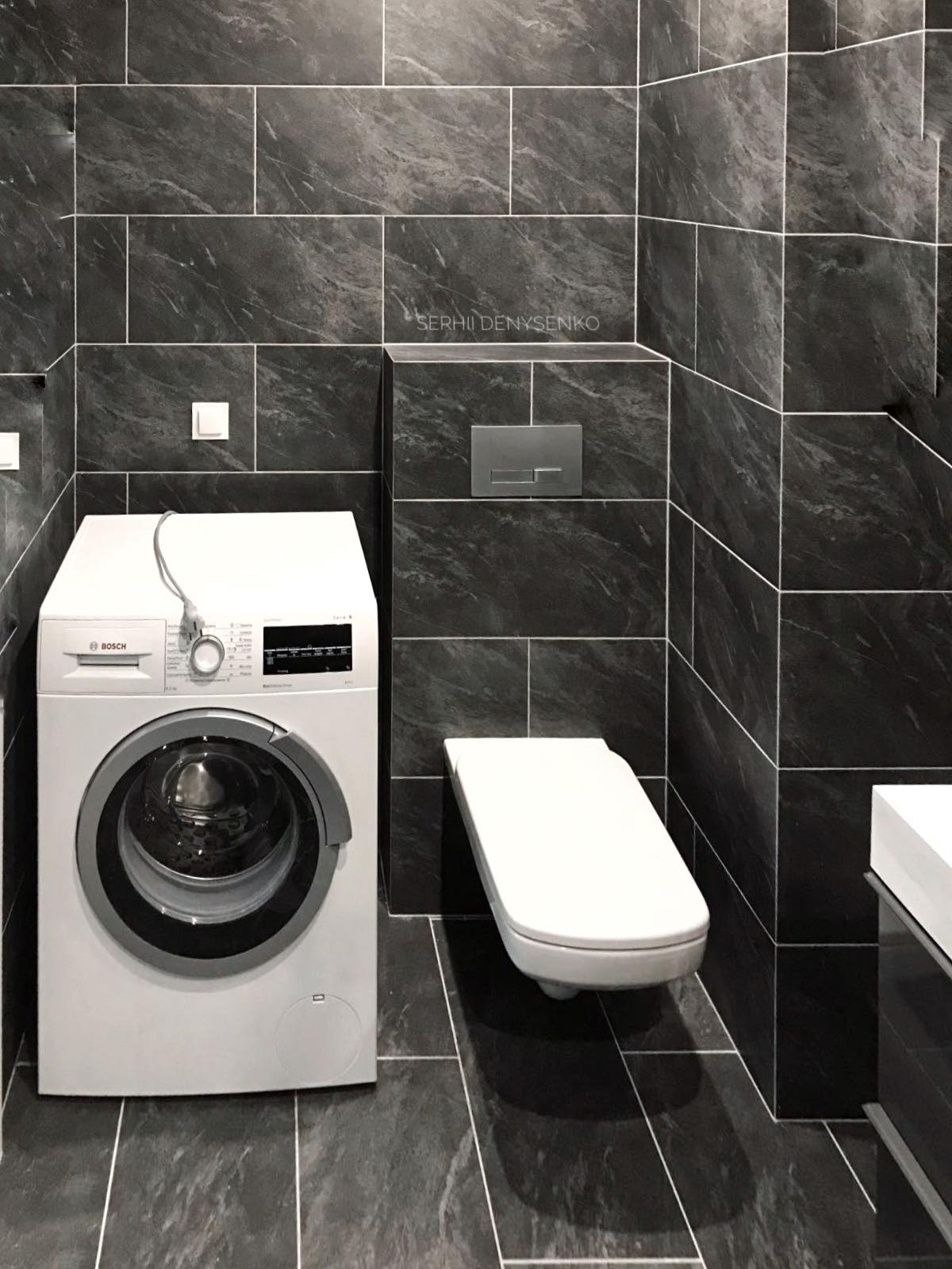 Nowoczesna łazienka z ciemnymi płytkami imitującymi marmur, wisząca toaleta, pralka Bosch z podłączonym kablem zasilającym i minimalistyczna armatura.