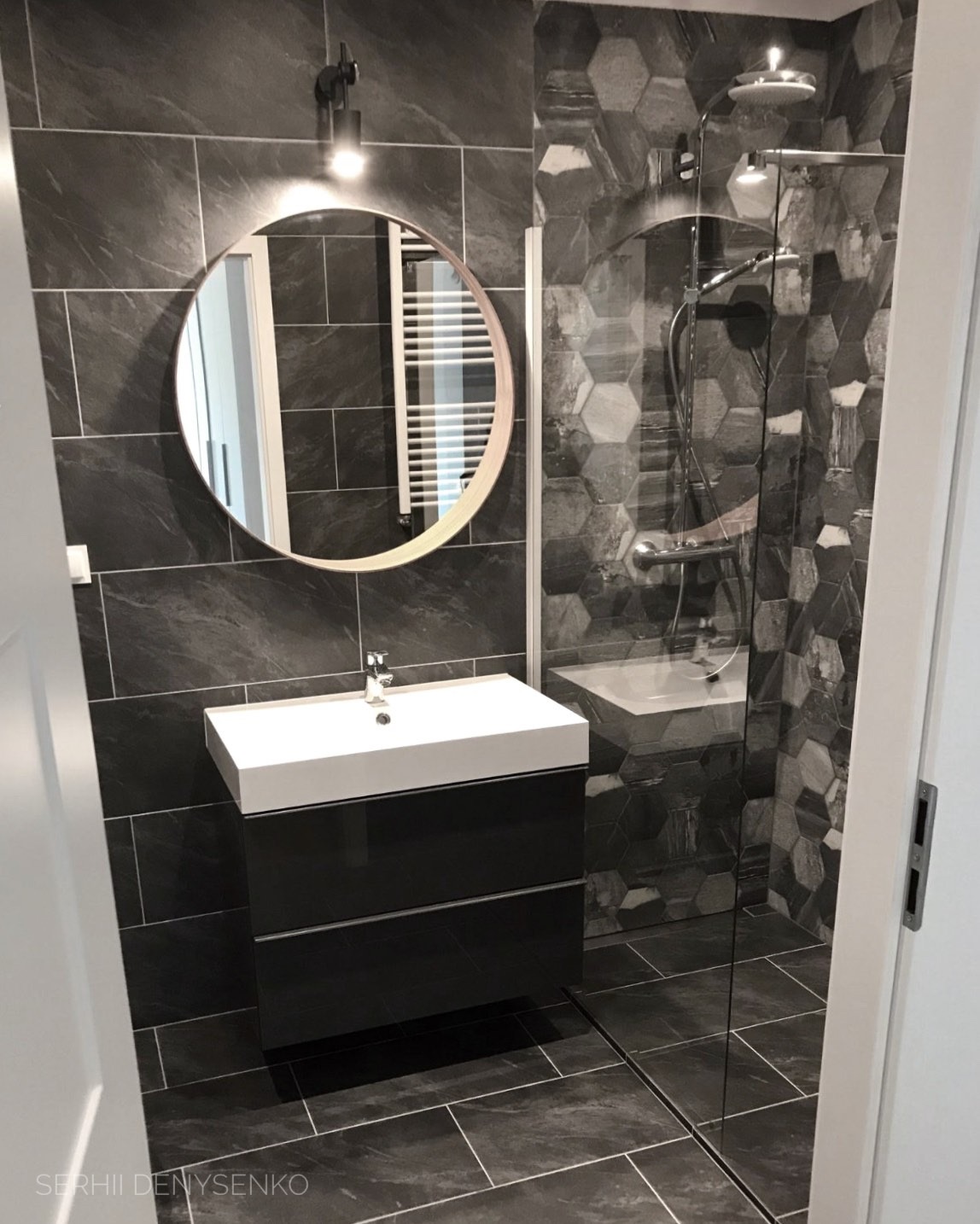 Nowoczesna łazienka w odcieniach szarości z heksagonalnymi płytkami pod prysznicem, okrągłe lustro z podświetleniem nad białą umywalką z czarną szafką, widok zza uchylonych drzwi.