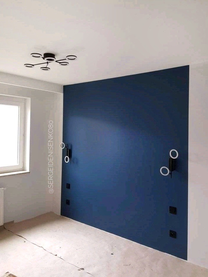 Ściana pomalowana na granatowo w pokoju z nowoczesnym oświetleniem ściennym i sufitowym, widoczne gniazdka elektryczne, surowa podłoga przed wykończeniem.