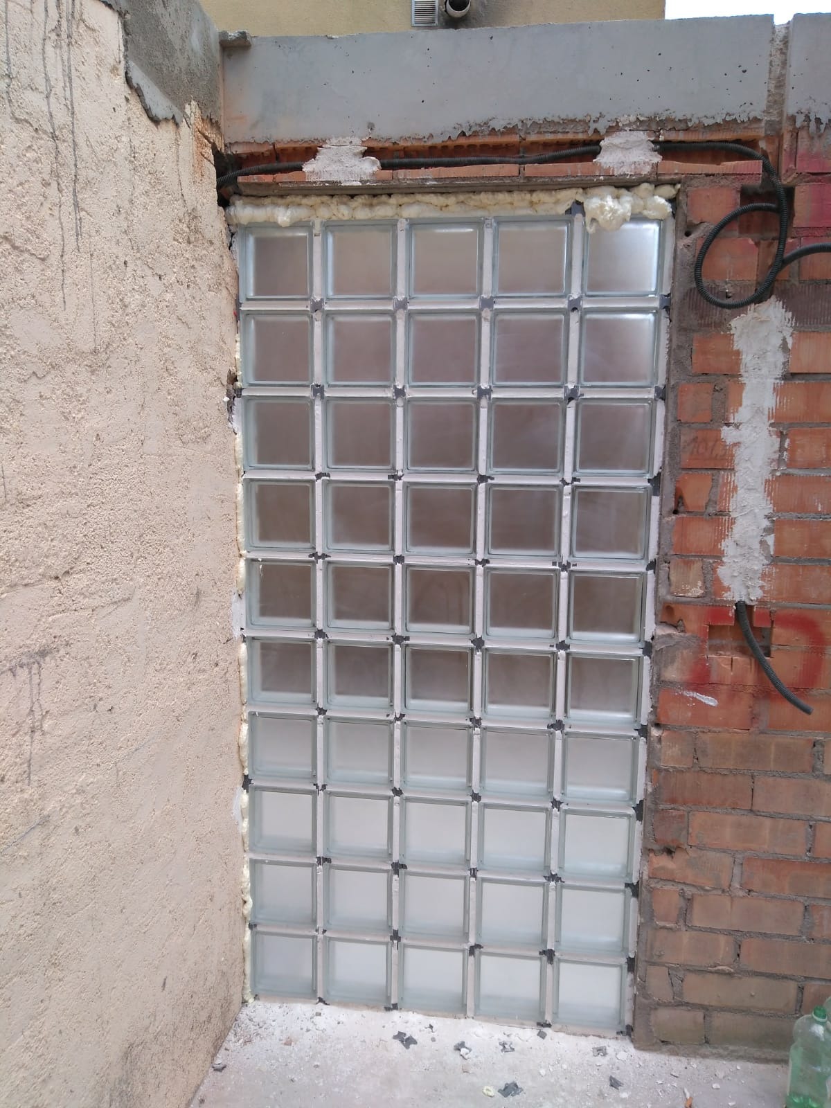 Ściana z luksferów w trakcie montażu, widoczna piana montażowa, surowy mur z cegły i fragment ściany z tynkiem.