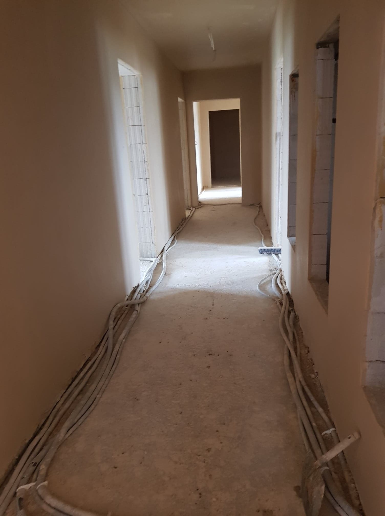 Korytarz w trakcie prac wykończeniowych: ściany pokryte świeżym, beżowym tynkiem, widoczne przewody elektryczne ułożone wzdłuż ściany, surowy beton na podłodze, brak listew przypodłogowych, otwory...