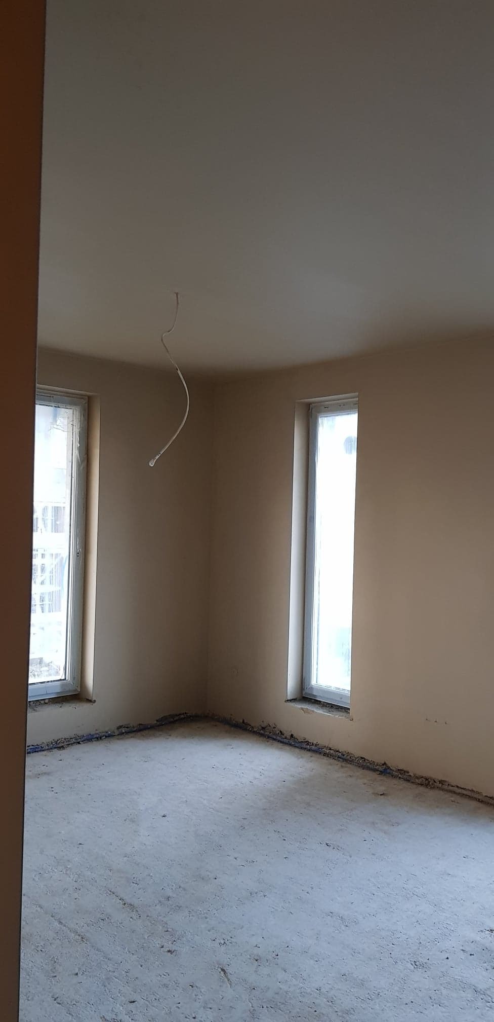 Pokój po nałożeniu jasnego tynku, widoczne dwa okna, wystający kabel ze ściany i resztki materiałów budowlanych na podłodze.