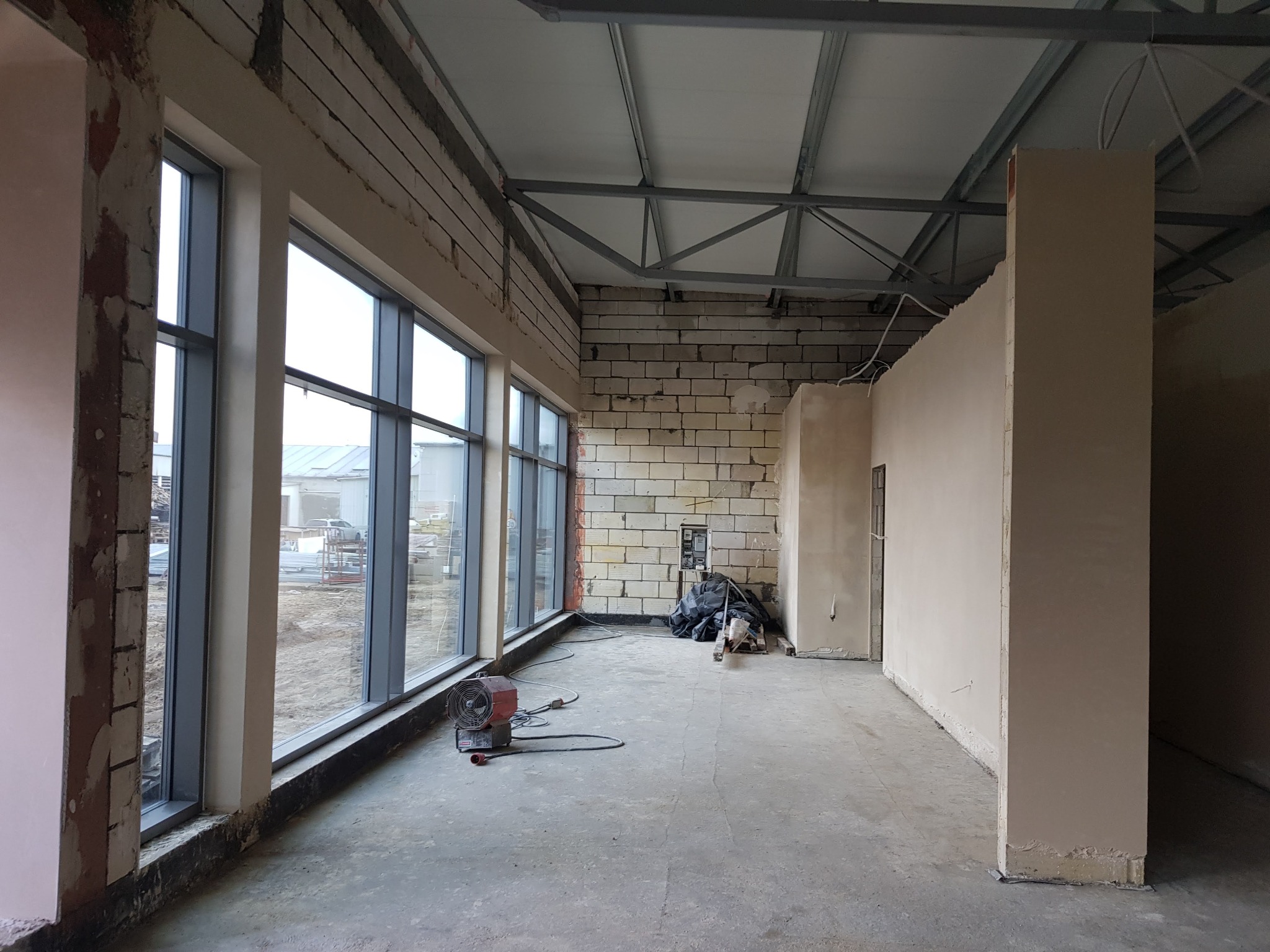 Wnętrze budynku w trakcie prac wykończeniowych: ściany z białej cegły, fragmenty ścian z nałożonym tynkiem, duże okna z widokiem na zewnątrz, metalowa konstrukcja dachu, nagrzewnica na podłodze.