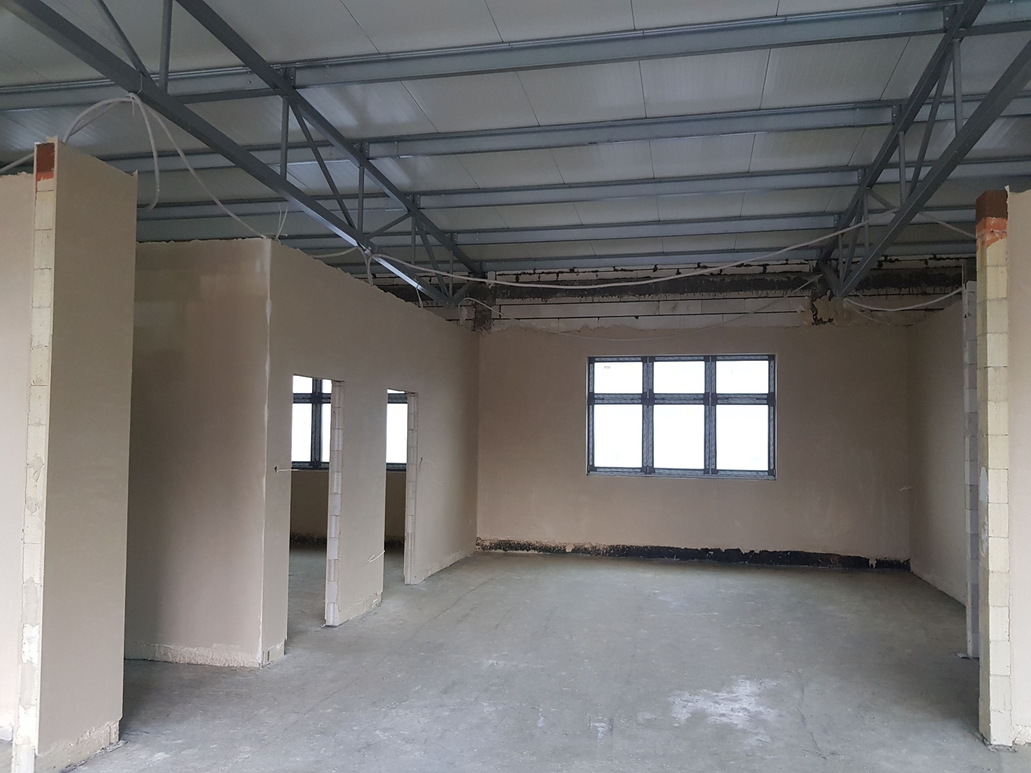 Surowe wnętrze budynku w trakcie prac wykończeniowych, ściany pokryte świeżym, jasnym tynkiem, widoczne okna i metalowa konstrukcja dachu.