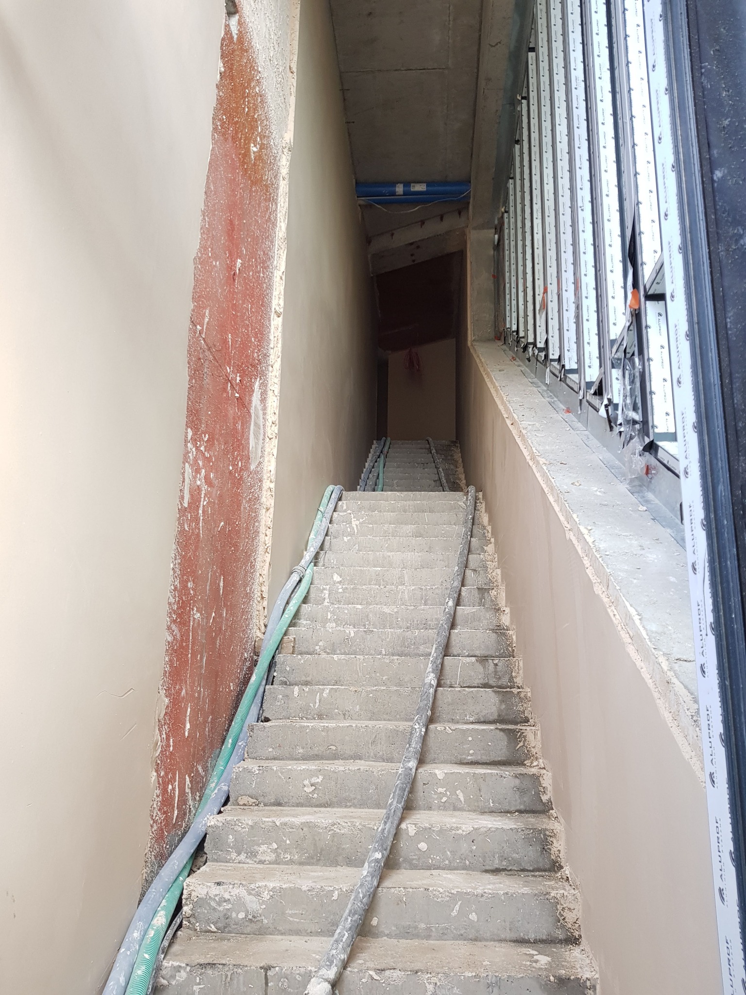 Klatka schodowa w trakcie prac tynkarskich; ściany częściowo pokryte świeżym, jasnym tynkiem, widoczne przewody i okna z profili aluminiowych.