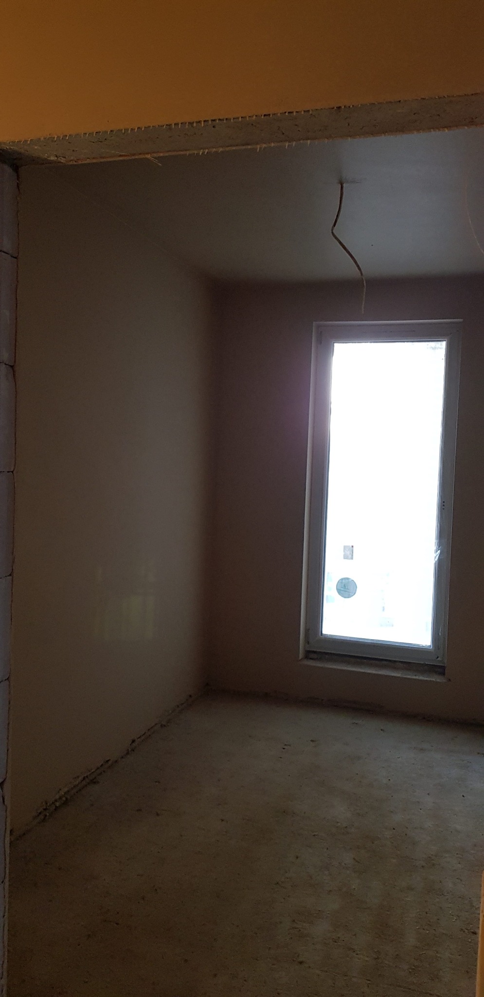 Pokój po nałożeniu gładzi gipsowej, widoczne świeżo otynkowane ściany i sufit, okno z intensywnym światłem dziennym wpadającym do wnętrza.