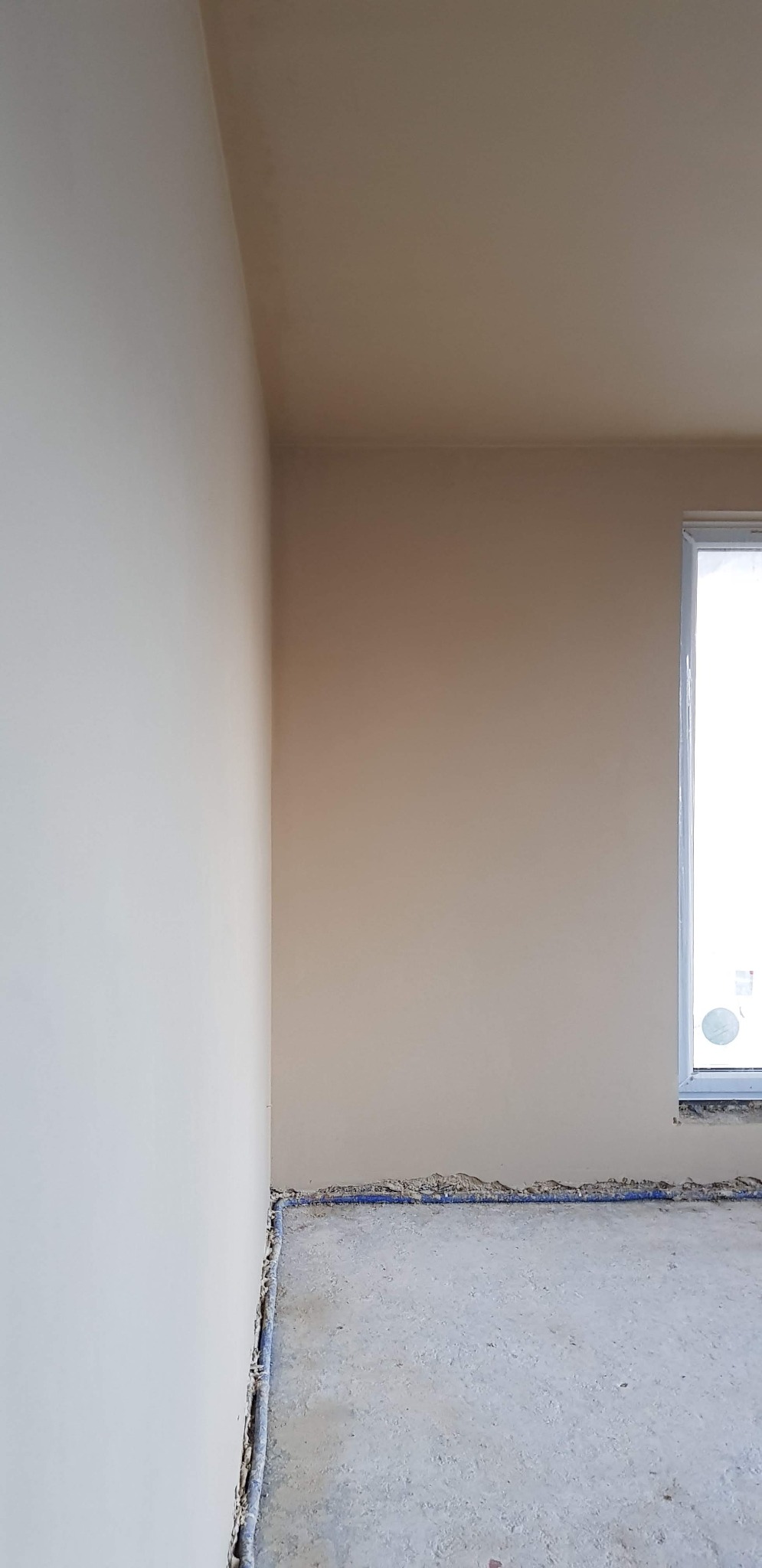 Wnętrze po nałożeniu gładzi, widoczne świeżo otynkowane ściany w odcieniach beżu i szarości, zarys okna w prawym górnym rogu oraz niezagospodarowana podłoga z wystającymi rurami wzdłuż ściany.