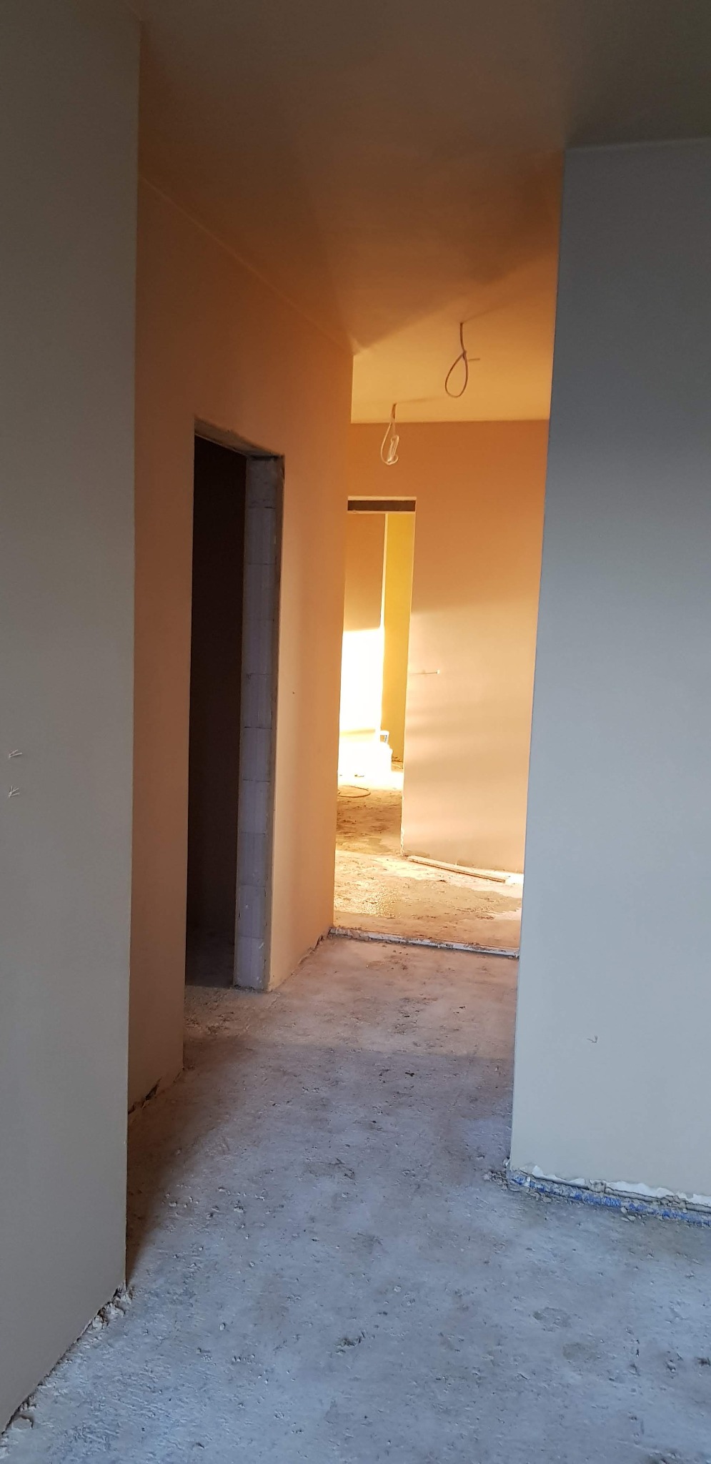 Korytarz w stanie surowym, ściany pokryte świeżym tynkiem, widoczne przewody elektryczne zwisające z sufitu, podłoga betonowa.