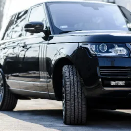 Czarny Range Rover z widoczną przednią oponą, chromowanym grillem i reflektorami LED, zaparkowany na betonowym podłożu z delikatnym rozmyciem tła.
