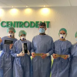 Zespół medyczny w uniformach i maseczkach pozuje z nagrodami na tle logo Centrodent, jedna osoba pokazuje kciuk w górę.